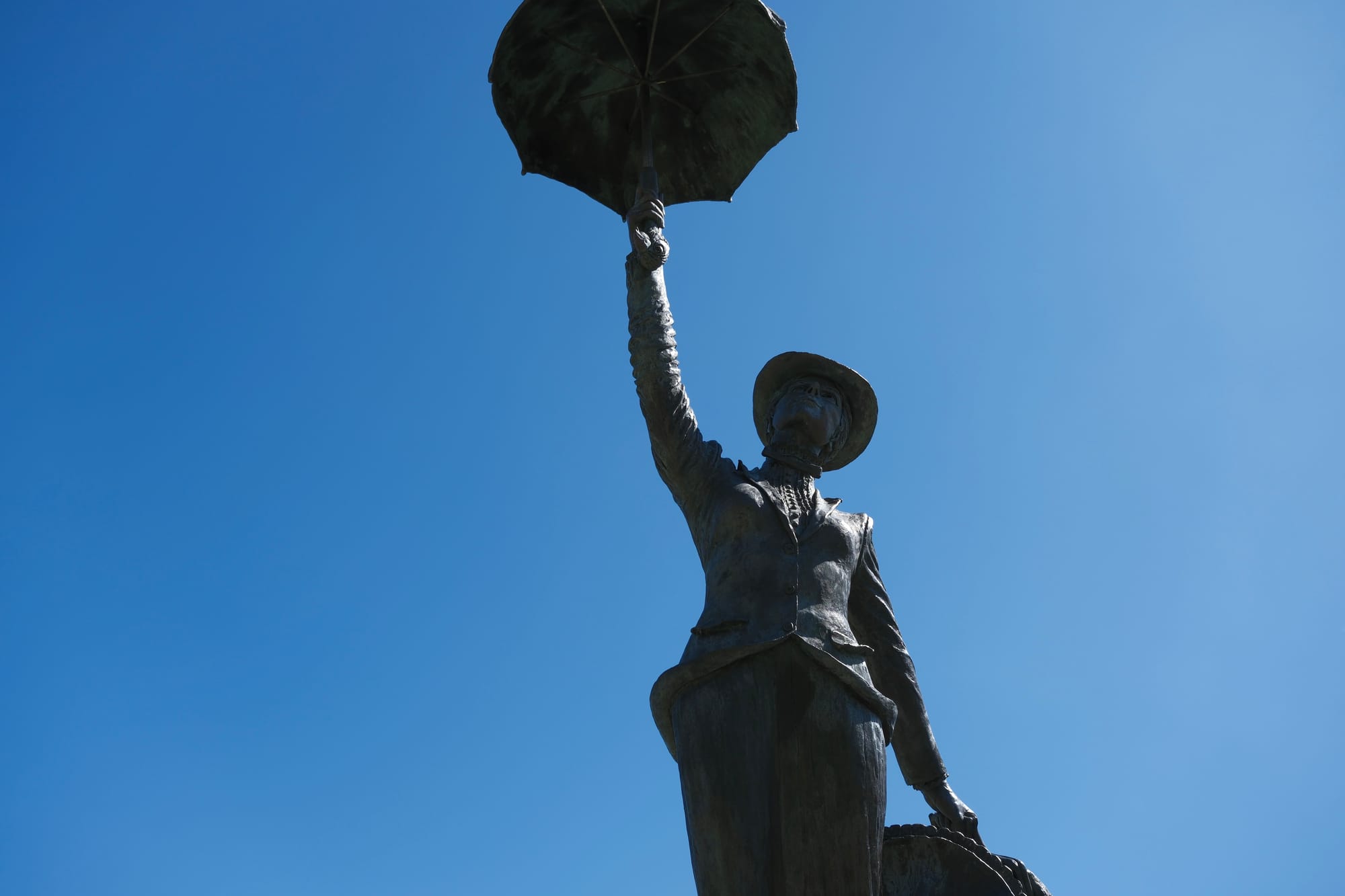 A bronze statue of Mary Poppins holding her umbrella high. Shot from below against a clear blue sky.