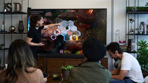 People in a living room with a large TV. One is standing pointing at a hex-grid map on the screen. The others are sitting and looking at phones as part of how they're playing.