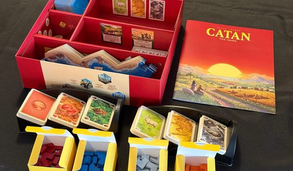 The open 6th Edition Catan box, with its cards, rules, and pieces organised on a black tablecloth.