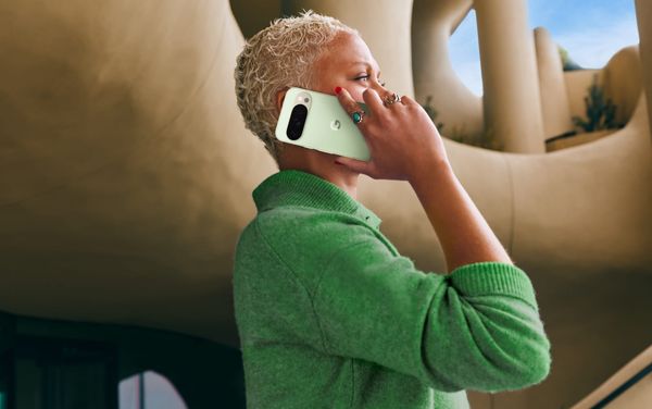 A woman holds a mint green Pixel phone to her ear, taking a call. She wears a green sweater and stands in front of a fancy artistic building.