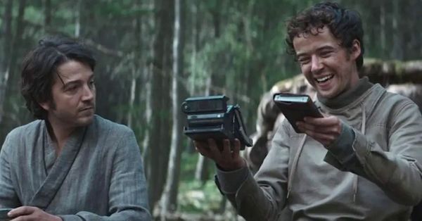 Two men sit in a forest. On the left, Andor looks with curiosity toward Nemik on the right. Nemik smiles as he holds up a notebook and an 'old' navigation device.