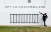Wall with iron fence with inscribed "MAY THESE GATES NEVER BE CLOSED". A white woman in black gestures at the words.