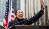 Alexandria Ocasio-Cortez, a youthful woman smiles and waves to a crowd at a lectern in front of an American f