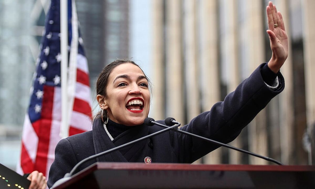 Alexandria Ocasio-Cortez, a youthful woman smiles and waves to a crowd at a lectern in front of an American f