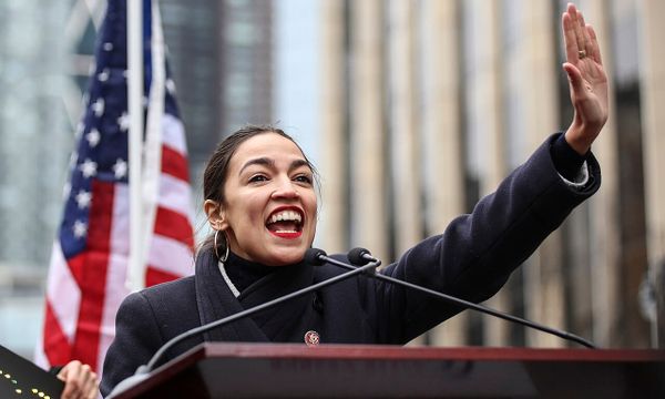 Alexandria Ocasio-Cortez, a youthful woman smiles and waves to a crowd at a lectern in front of an American f