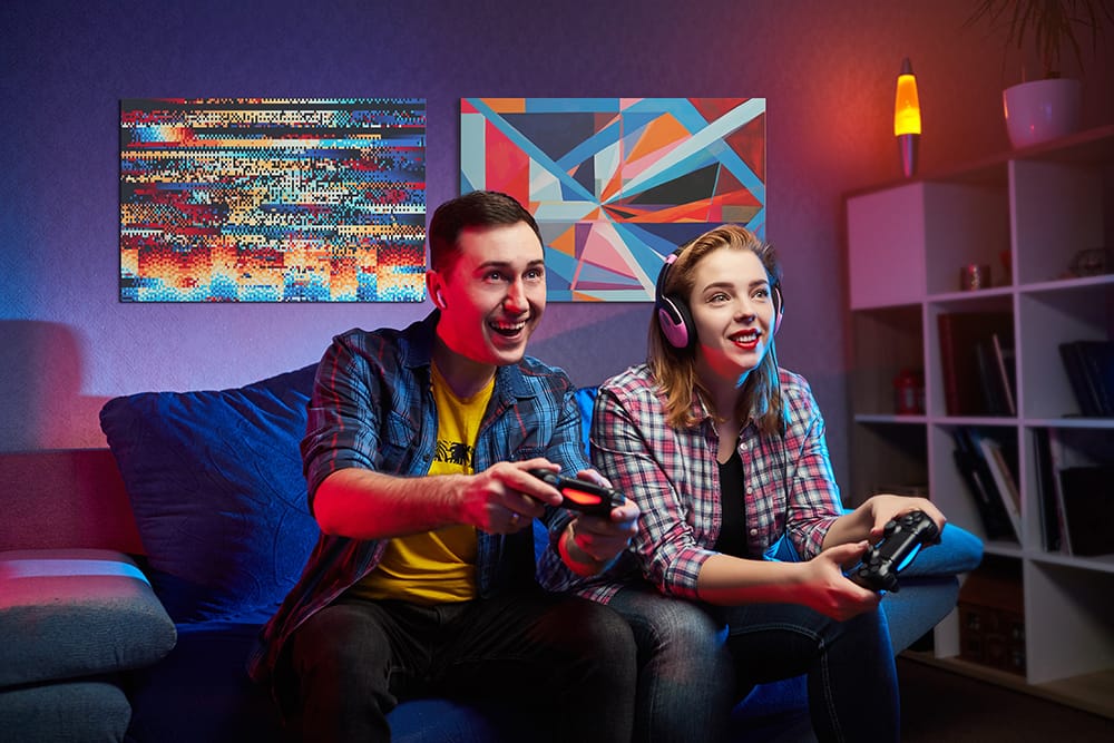 Two people playing video games on a couch with colorful abstract wall art and neon lighting.