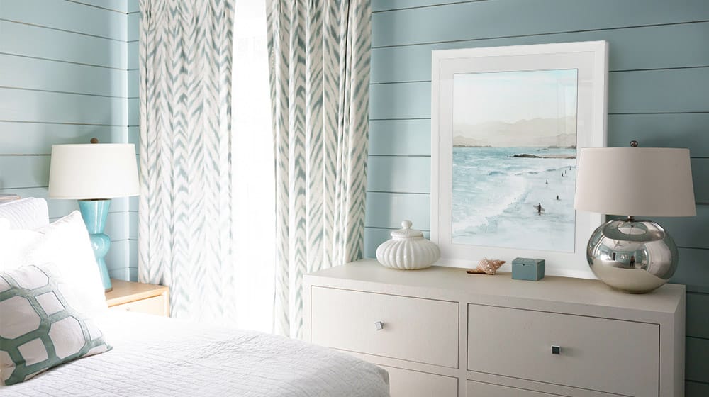 Coastal art in a white framed print leaning on a dresser in a coastal bedroom