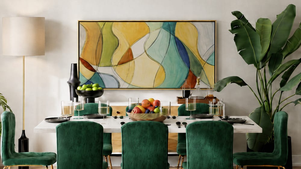 Gold floating framed canvas print with muted abstract art above a modern dining table with green velvet chairs, fruit centerpiece and indoor plants.