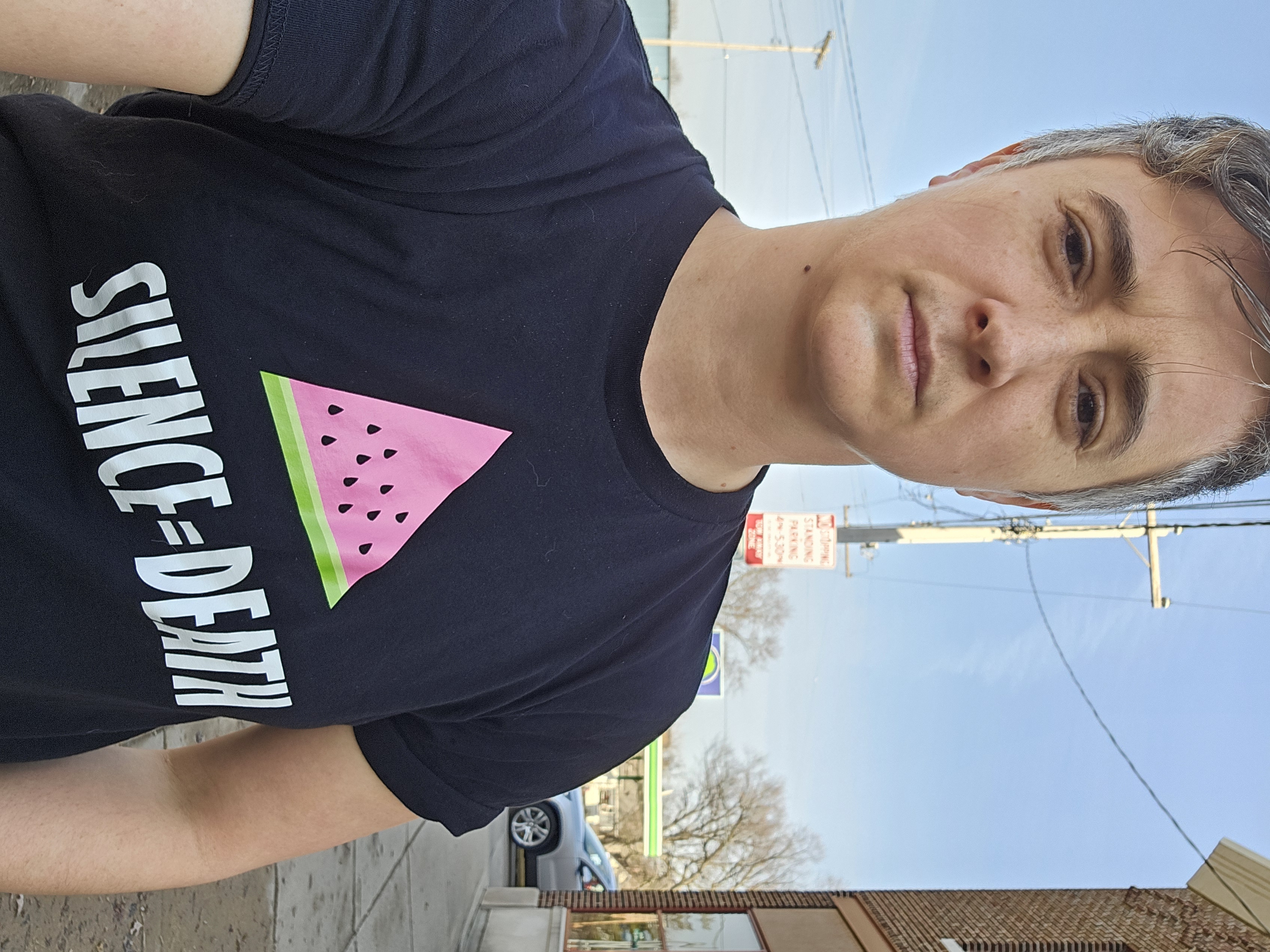 Emily looking into the camera, looking solemn, wears a black t-shirt with the image of a watermelon slice in the shape of the traditional gay pink triangle, and the words "Silence = Death" written below.