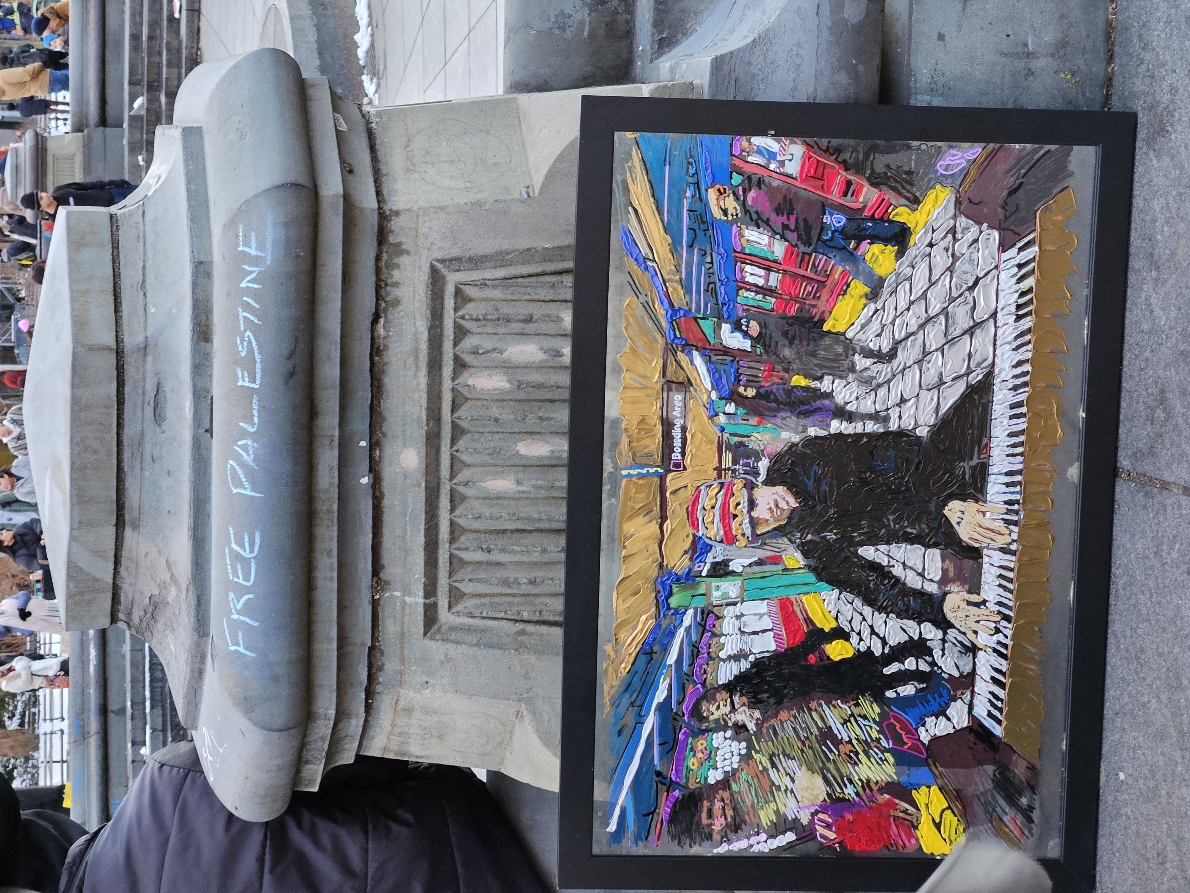 "Free Palestine" is written in blue chalk across the top of a cement pillar. A painting is leaned against the pillar that shows a colorful depiction of a pianist playing on a subway platform. 