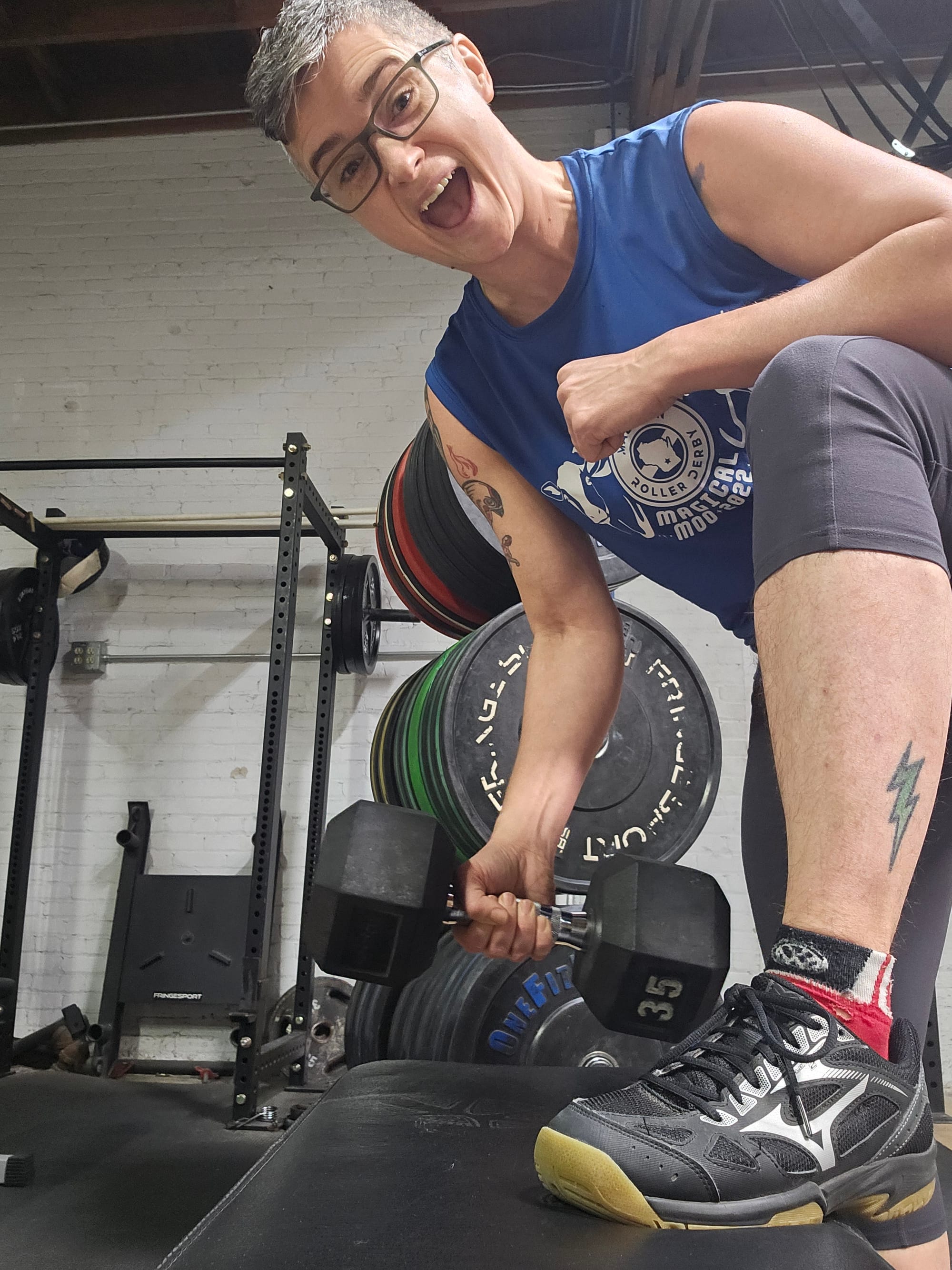 Emily poses with a large barbell in one hand and the opposite foot propped on top of a weight bench. They are smiling like a weirdo.
