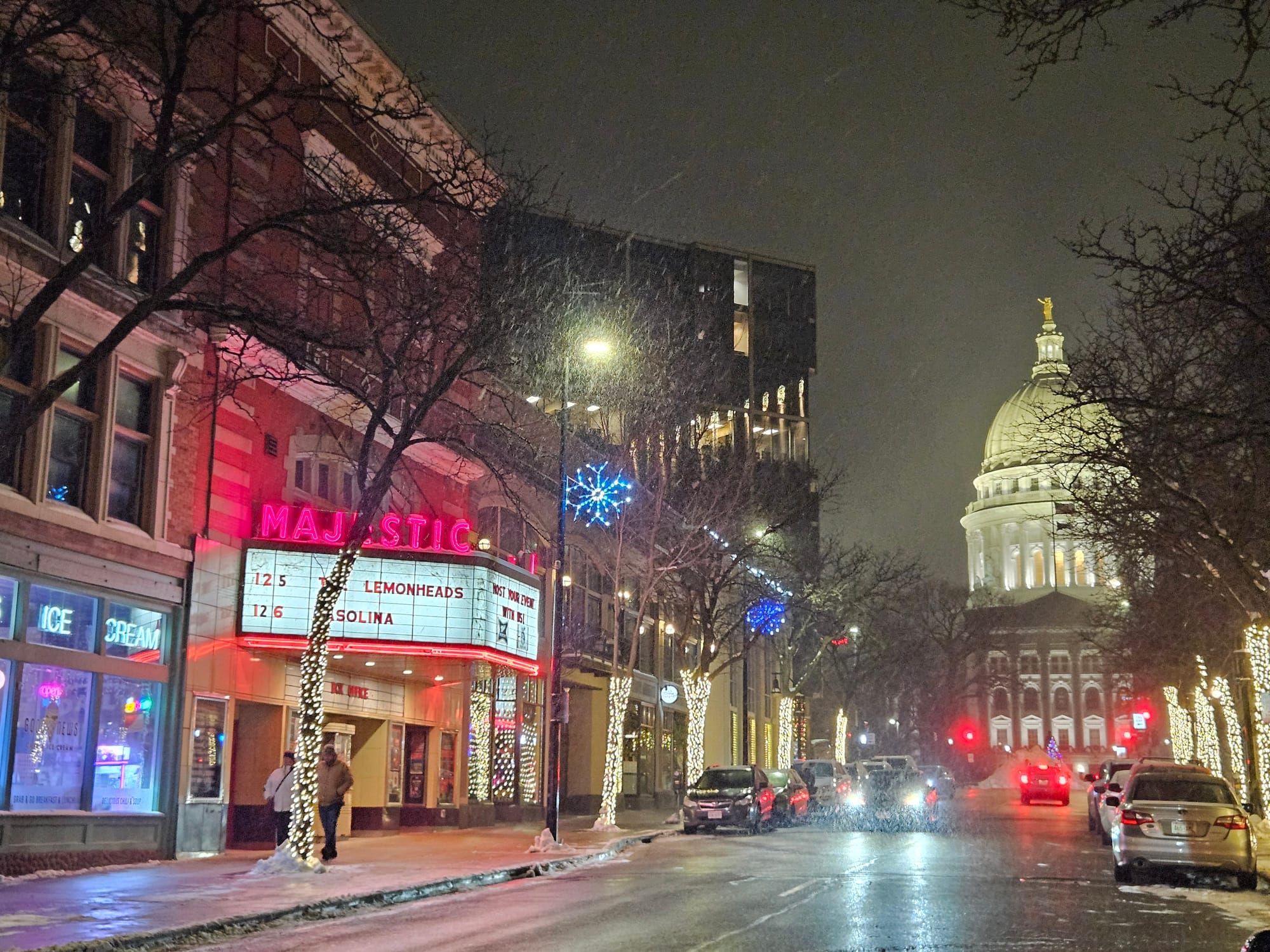 Snow falls on a nighttime street scene. At left, the Majestic Theater’s lit-up marquee, and at right, the tall white dome of the Wisconsin State Capitol.