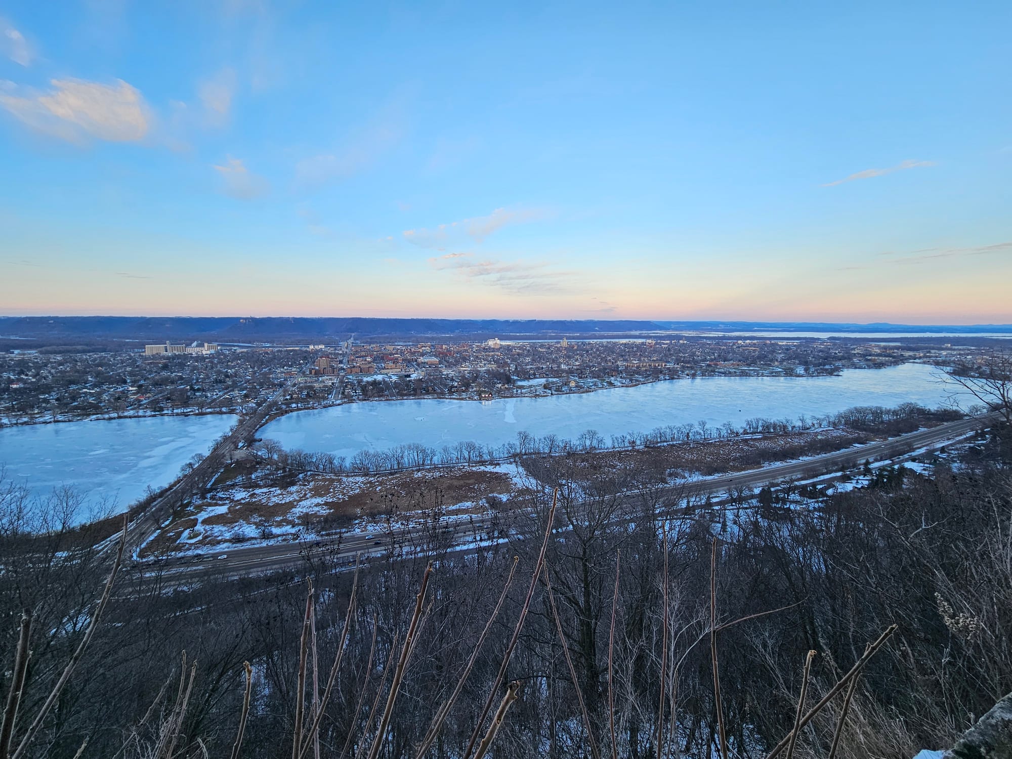 A view from the side of a bluff shows a long icy lake bordering a sprawling town, with a river and more bluffs visible in the distance.