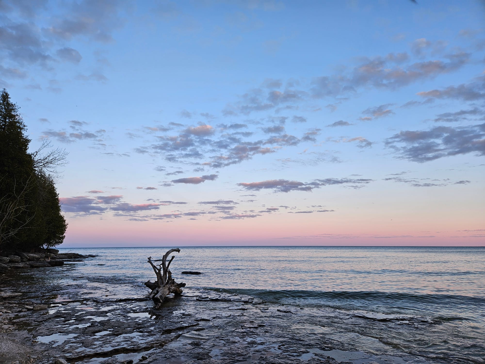 Waves lap a rocky shoreline, with the pink and purple light of a sunset on the horizon and blue sky overhead.