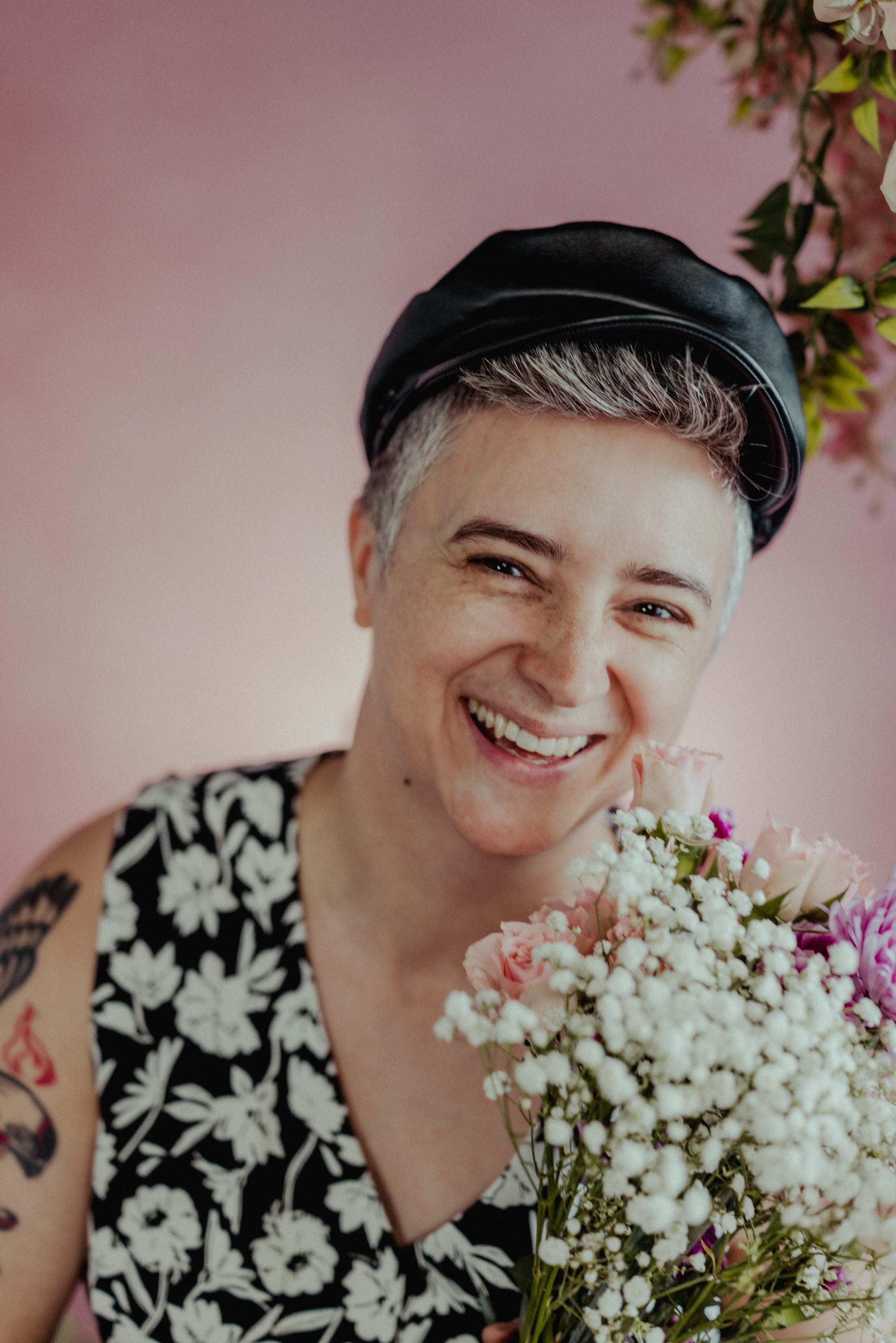 Emily, a white, masc-leaning person, smiles broadly into the camera while holding a bouquet of flowers. They are wearing a black leather hat and a black vest with a white floral pattern.