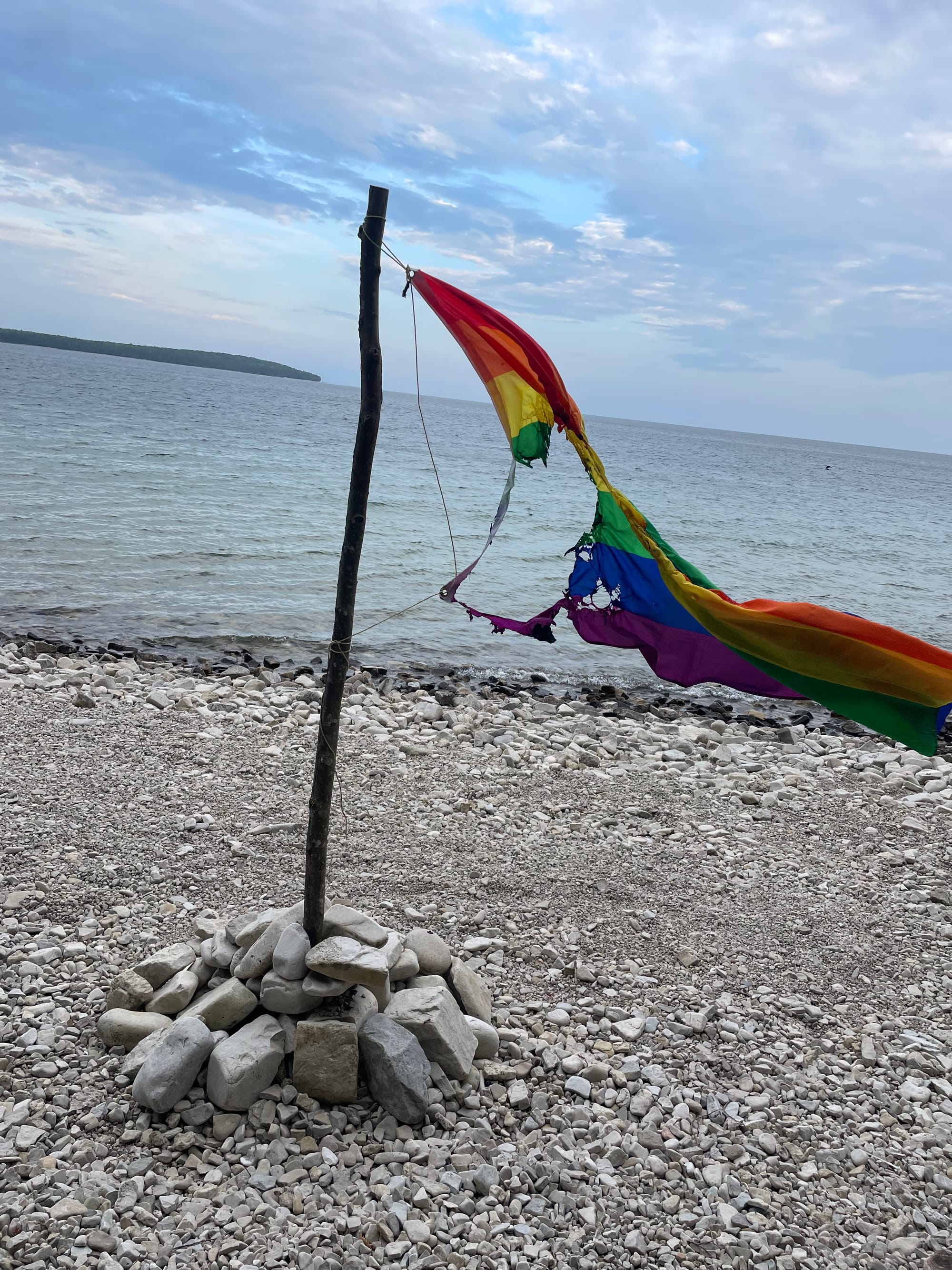 A rainbow Pride flag, still attached to the wooden branch used as a flagpole, with a huge hole burned into its center.