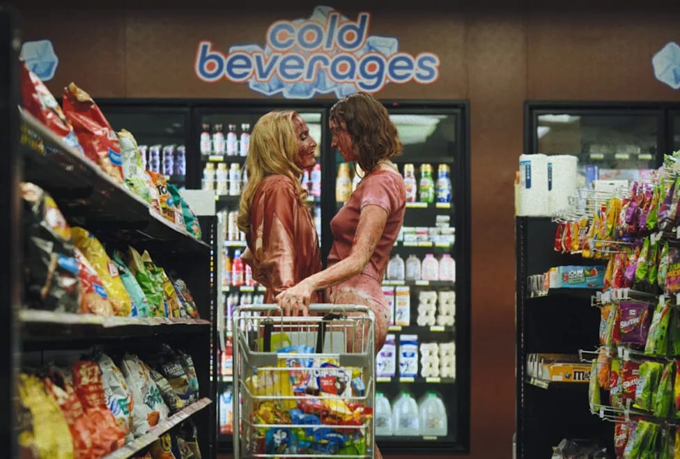 Two women, both covered in blood, standing holding hands over a shopping cart full of candy, in the aisle of a convenience store.