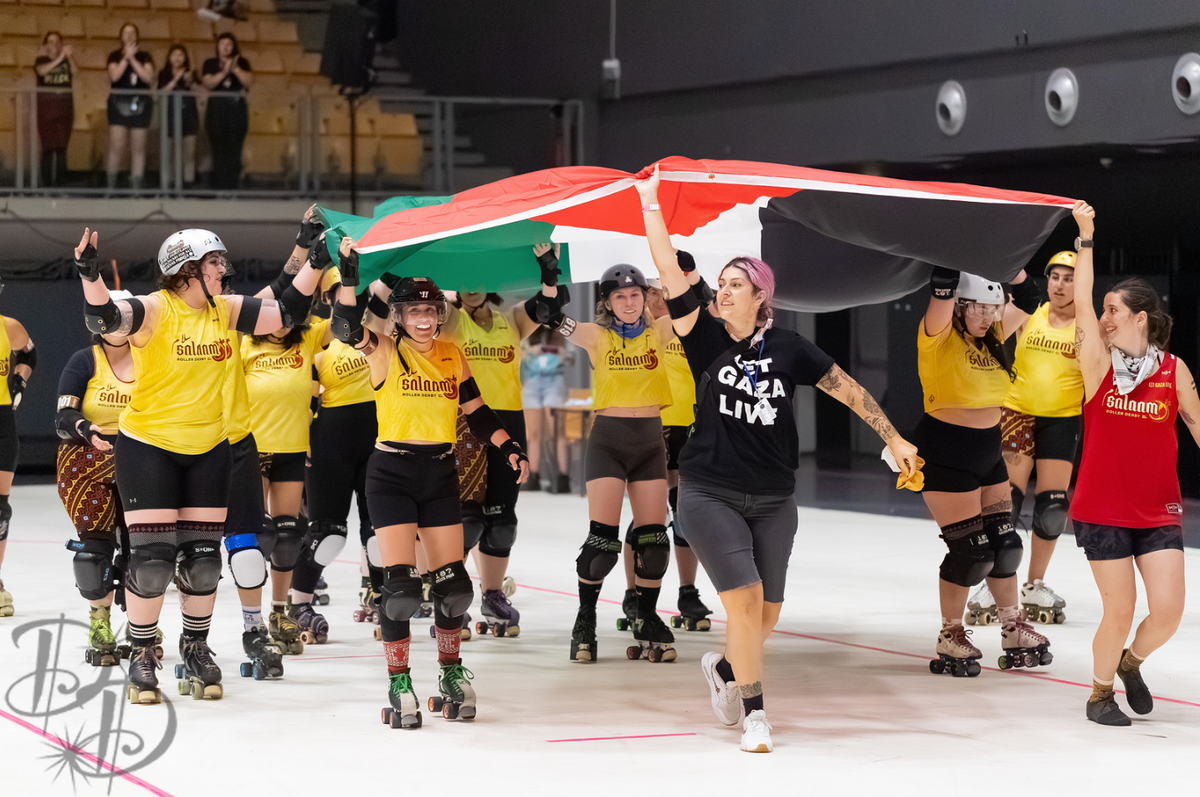 A group of roller derby skaters and coaches carry a large Palestinian flag overhead as they skate and walk in a procession.