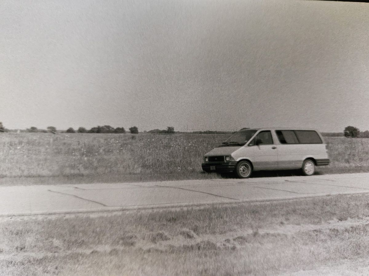 A black and white photo shows an old Ford minivan parked along a cracked roadway, with a flat field of grass and flowers behind it.