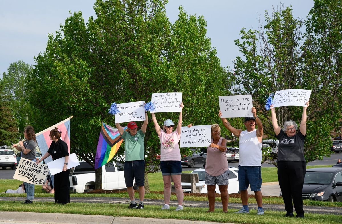 A group of people stands along a roadside holding signs in the air that hold messages of support for music and LGBTQ+ rights.
