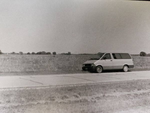 A black and white photo shows an old Ford minivan parked along a cracked roadway, with a flat field of grass and flowers behind it.