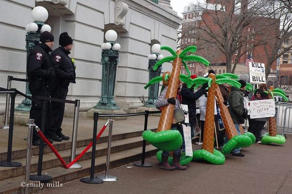 Two cops stand watch over a line of protesters holding giant inflatable palm trees outside the Wisconsin State Capitol building in winter.