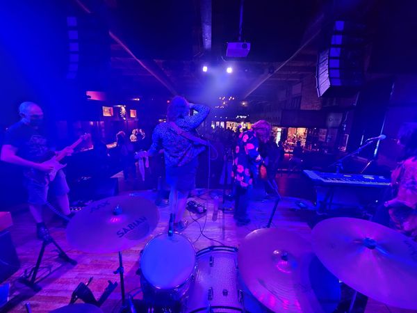 A view looking out over a drum set, with band members on stage in front, speakers overhead, and a crowd beyond.