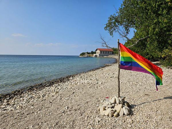 A rainbow Pride flag flaps in the breeze along a rocky shore.