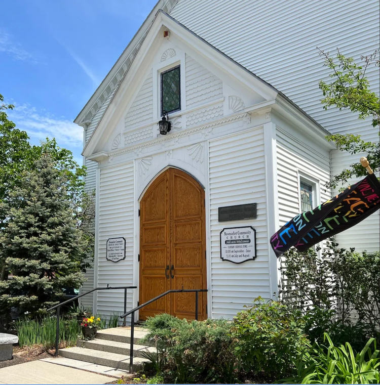 Steps lead up to the oak, arched doors of a white clapboard church. A small garden surrounds the steps. A multicolored banner announcing peace and acceptance of all flutters in the breeze.   