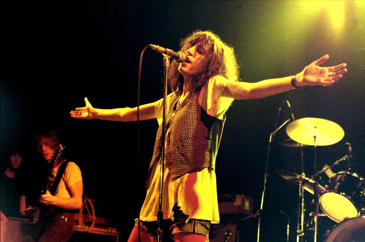 Patti Smith on stage, with eyes closed, singing with arms outstretched as if in prayer.