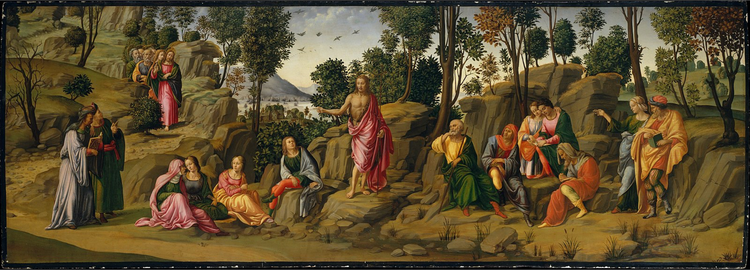 A man in red robes stands on a rock to address a crowd. Water and trees are in the background.