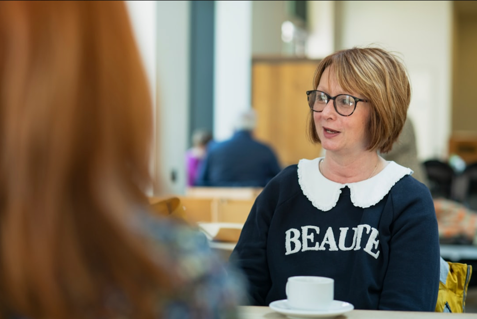 A woman with dark blonde hair, glasses, and a dark blue sweater with "BEAUTE" on it. She speaks with other women at her table. 