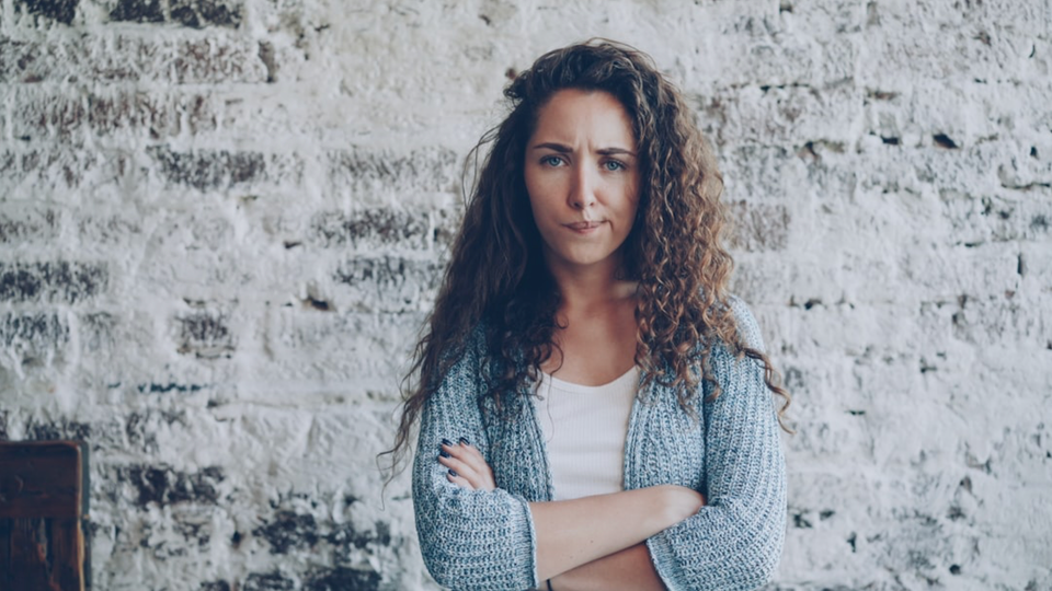 A young woman with brown curly hair stares at the viewer, looking disappointed. Her arms are crossed.