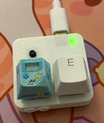 A 3D printed square holds two keyboard keys: one that looks like a tiny turquoise gameboy and the other the letter 'E'
