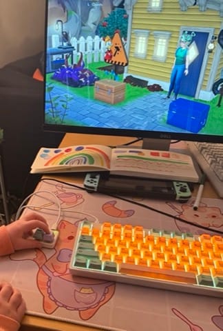 A child's hand hovers over a two-key keyboard while an illuminated full keyboard sits to its right. In the background, a computer monitor shows Disney Dreamlight valley