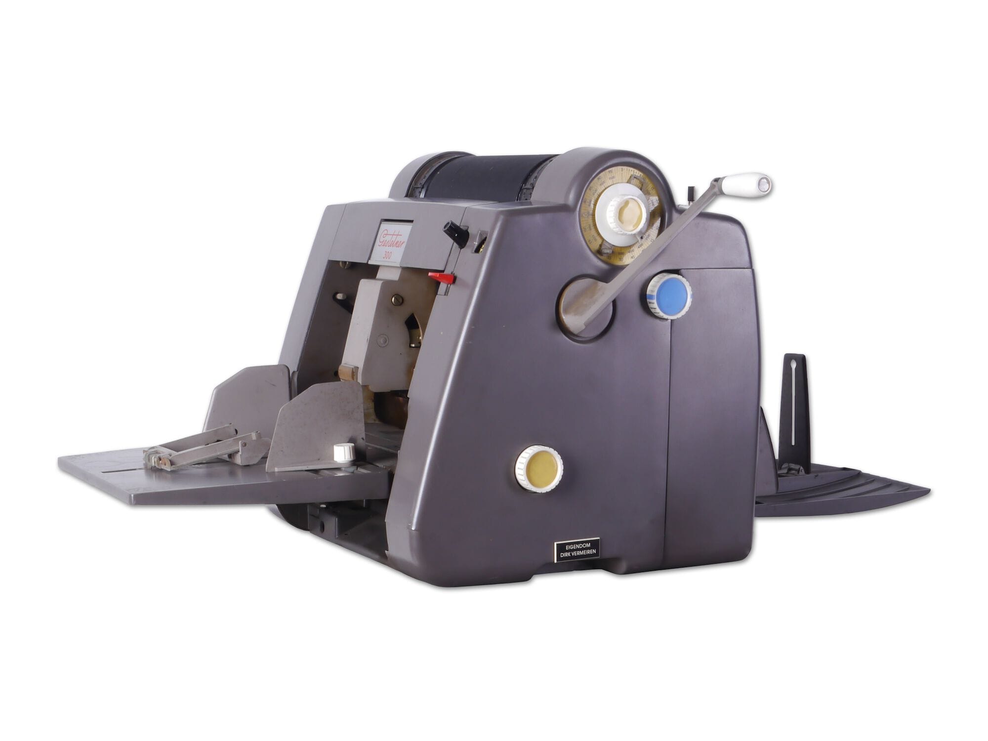 Gestetner, type 300, mimeograph machine. Grey, solid. It has a feed tray and a receiving tray for paper. There is a handle that turns the ink cylinder on the top.