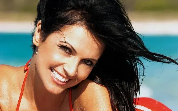 Denise Milani smiling in swimwear with an American flag behind her.