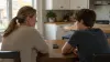 A parent and teenage child discuss smartphone use at a kitchen table in natural afternoon light.