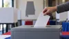 A voter places an unmarked ballot into a secure ballot box with a subtle Alabama silhouette in the background.