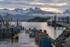 An Alaska fishing harbor and ferry dock with workers, equipment and snow-dusted mountains under a cloudy morning sky