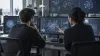 Two technical evaluators review abstract AI system tests on monitors in an unbranded laboratory.