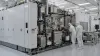 Two semiconductor cleanroom technicians inspect a large unbranded advanced lithography system.