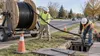 Telecommunications technicians guide fiber-optic cable from a large reel into an underground conduit.