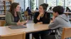 Teacher, parent and student discuss school technology beside a closed laptop in a sunlit school library.