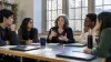 A professor and four college students discussing ideas around a seminar table in a sunlit classroom.