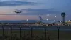 A small drone flies near the perimeter of a generic airport at dusk as an unbranded passenger jet waits near a control tower.