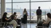 Travelers wait beside carry-on bags in a sunlit airport departure area overlooking an unbranded passenger jet.