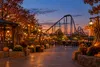 Illustrative fall festival promenade at a Texas amusement park with pumpkins, string lights and a roller coaster at dusk.