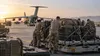 Service members load unmarked cargo pallets near a transport aircraft at a desert airfield.