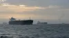 Commercial oil tankers cross choppy water under a muted dawn sky with a distant naval vessel on the horizon.